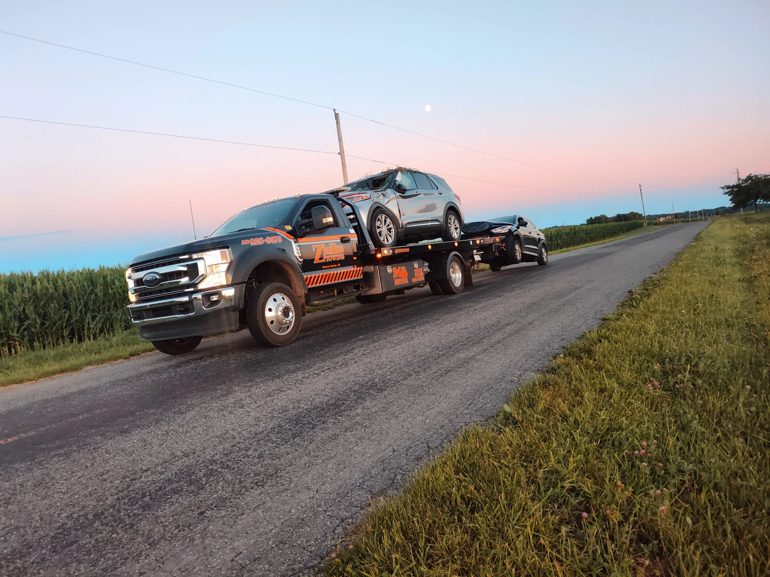 Zielke Towing light duty flatbed truck hauling two cars during dusk with a pink sky background