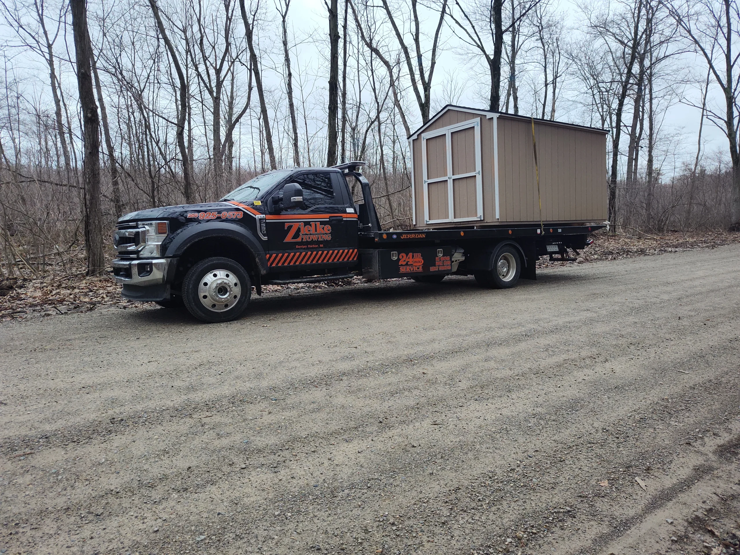 Zielke Towing light duty flatbed hauling a small shed on a gravel road surrounded by leafless trees