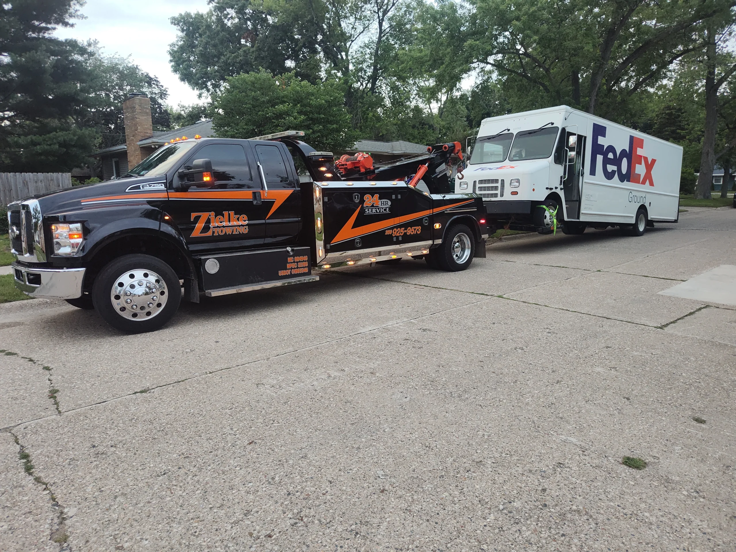 Zielke Towing flatbed truck hooked up to a broken-down FedEx box truck on a residential street